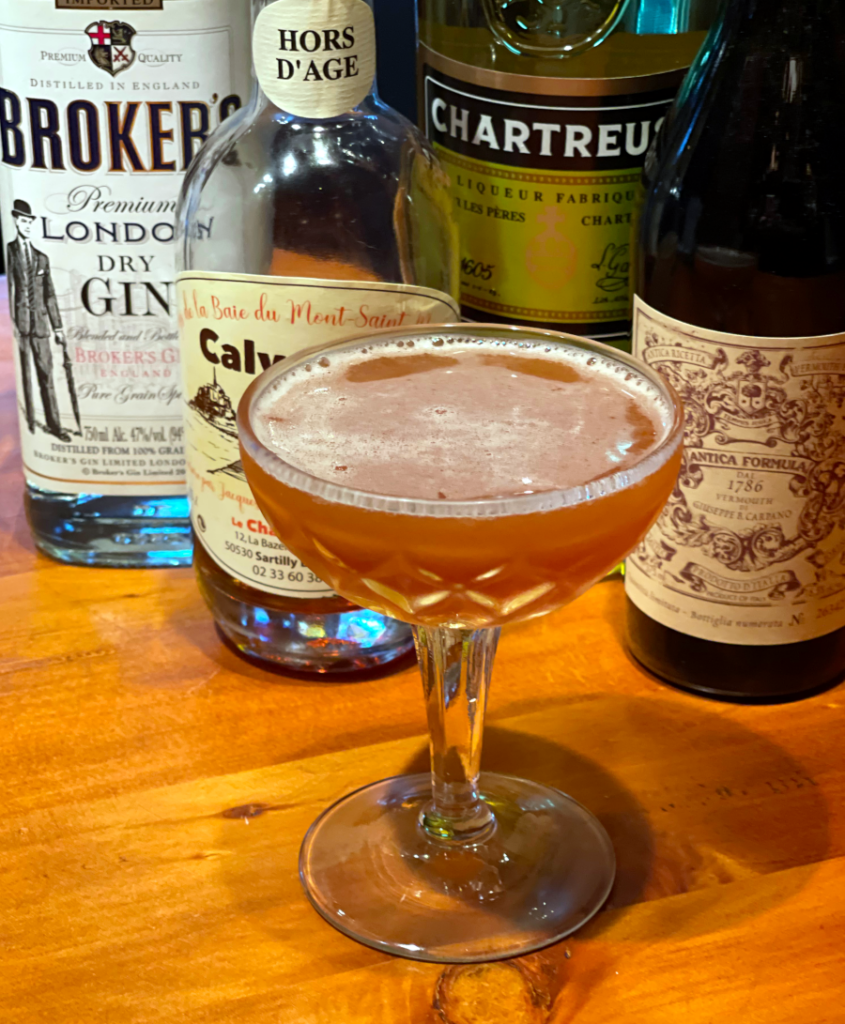 Peace Train Cocktail: a golden cocktail in a coupe glass in the foreground with ingredient bottles in the background